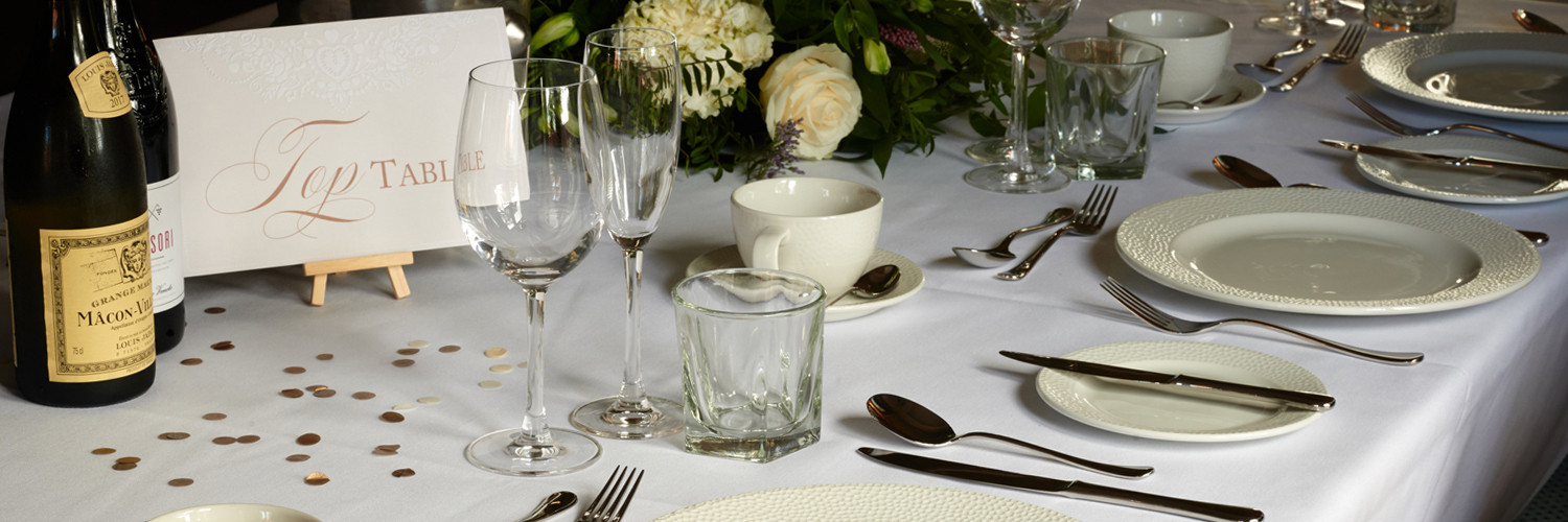 A wedding top table dressed with white table cloth, wine glasses and a place setting.